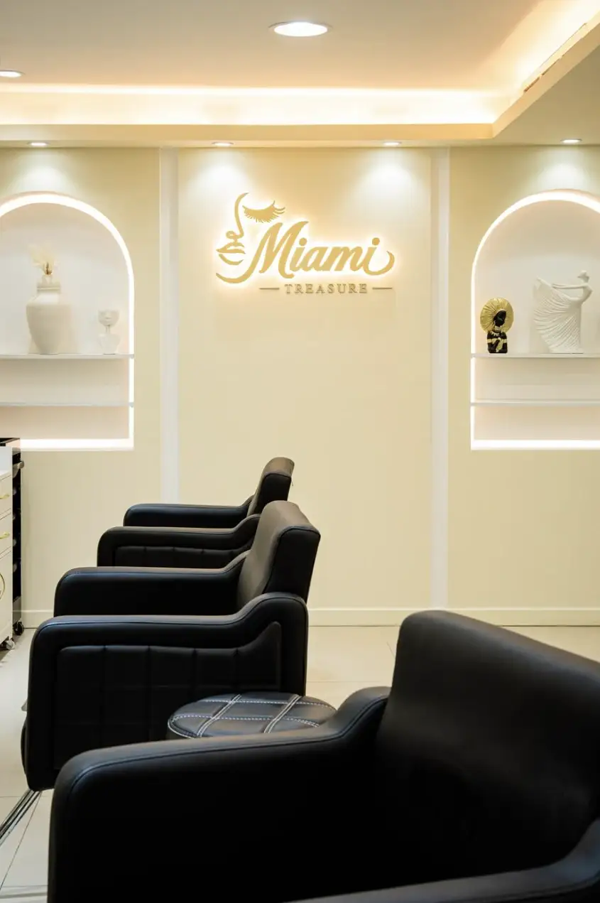 Two arched wall niches with shelves on either side of the gold-lit Miami Treasure logo, above a row of black styling chairs